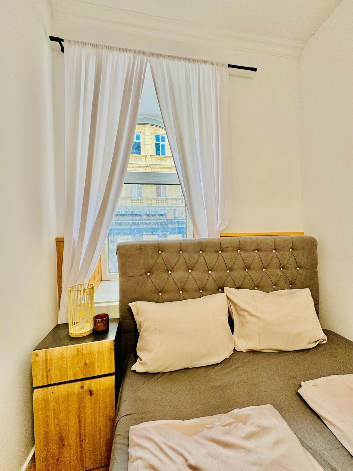 A bedroom with a bed, a wooden nightstand, a window with white curtains, and a view of a building outside.