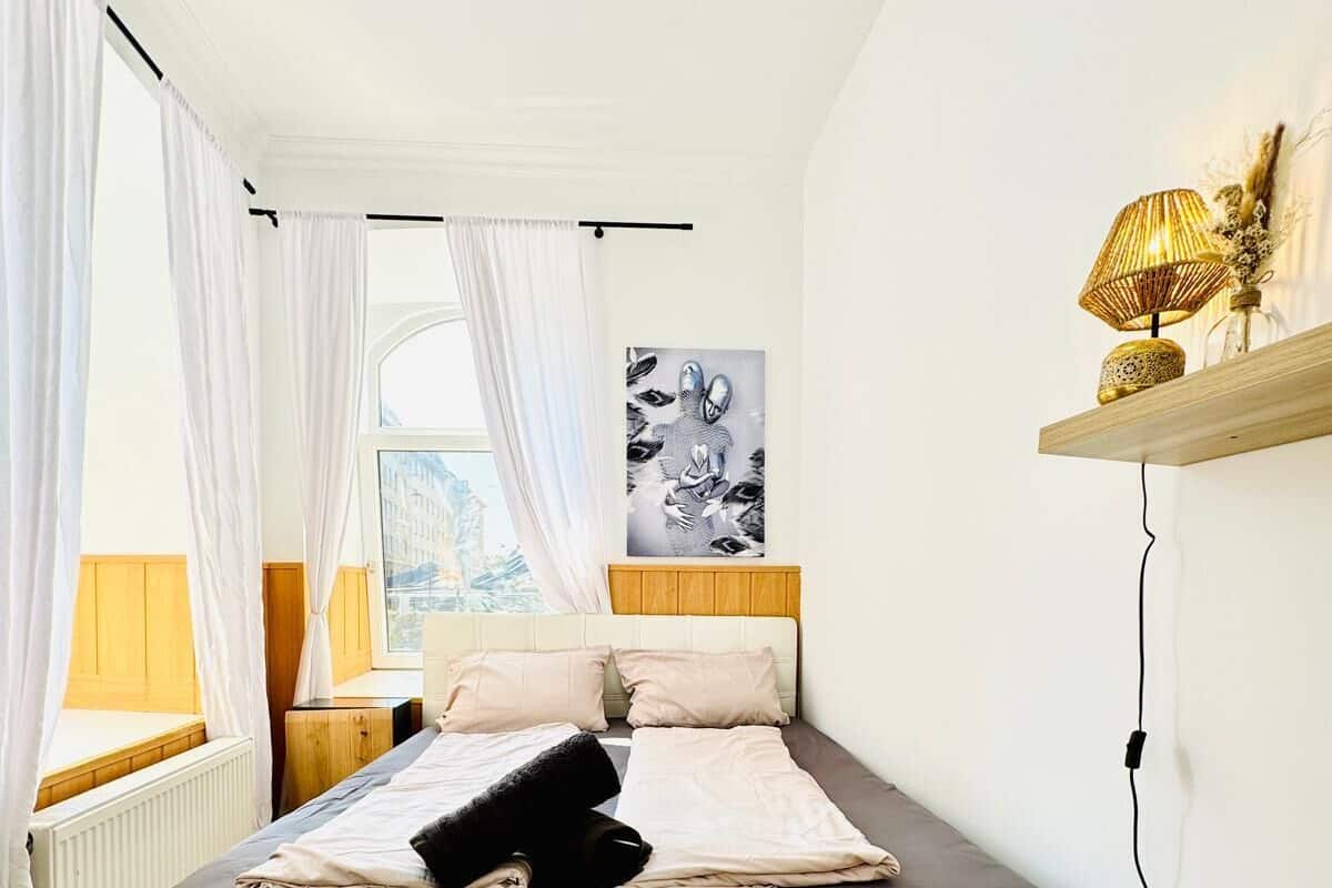 A modern bedroom with a bed, white curtains, a window, a wall-mounted shelf with decorative items, and a painting.