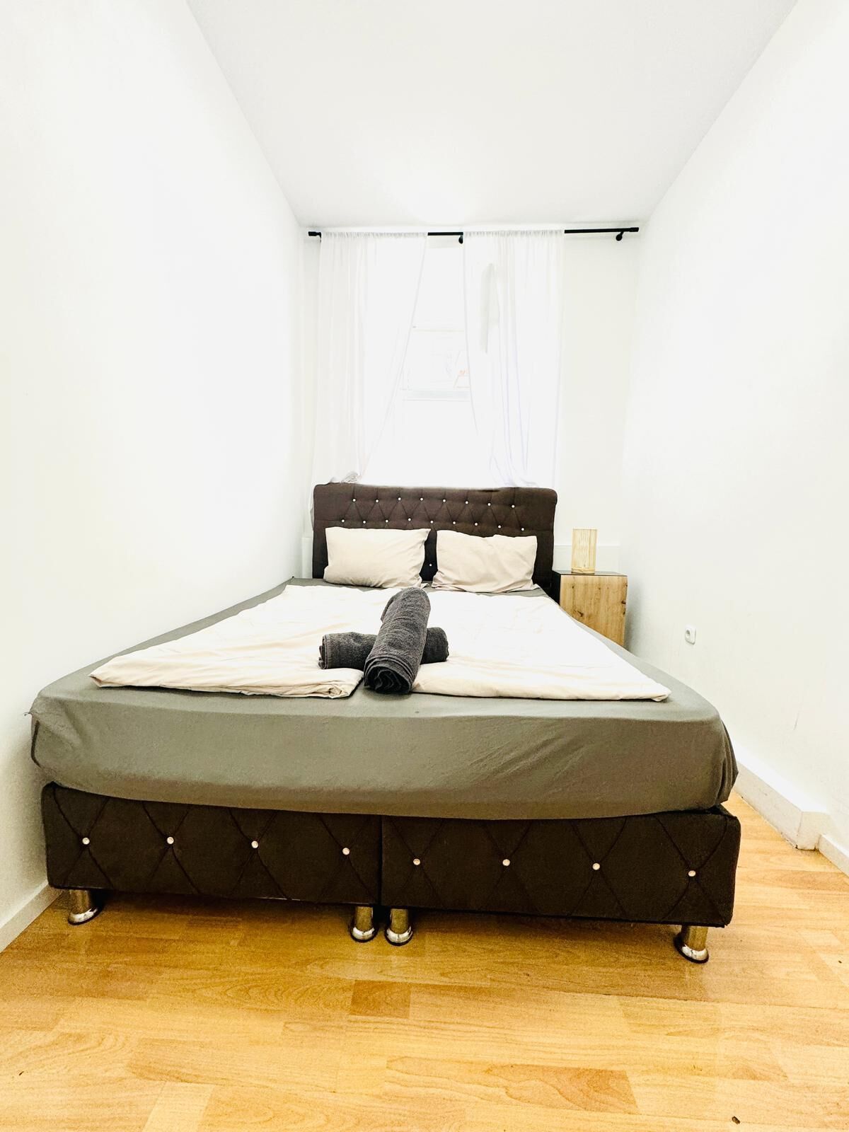 A single bed with a dark headboard and footboard, a wooden nightstand, and a window with sheer curtains.