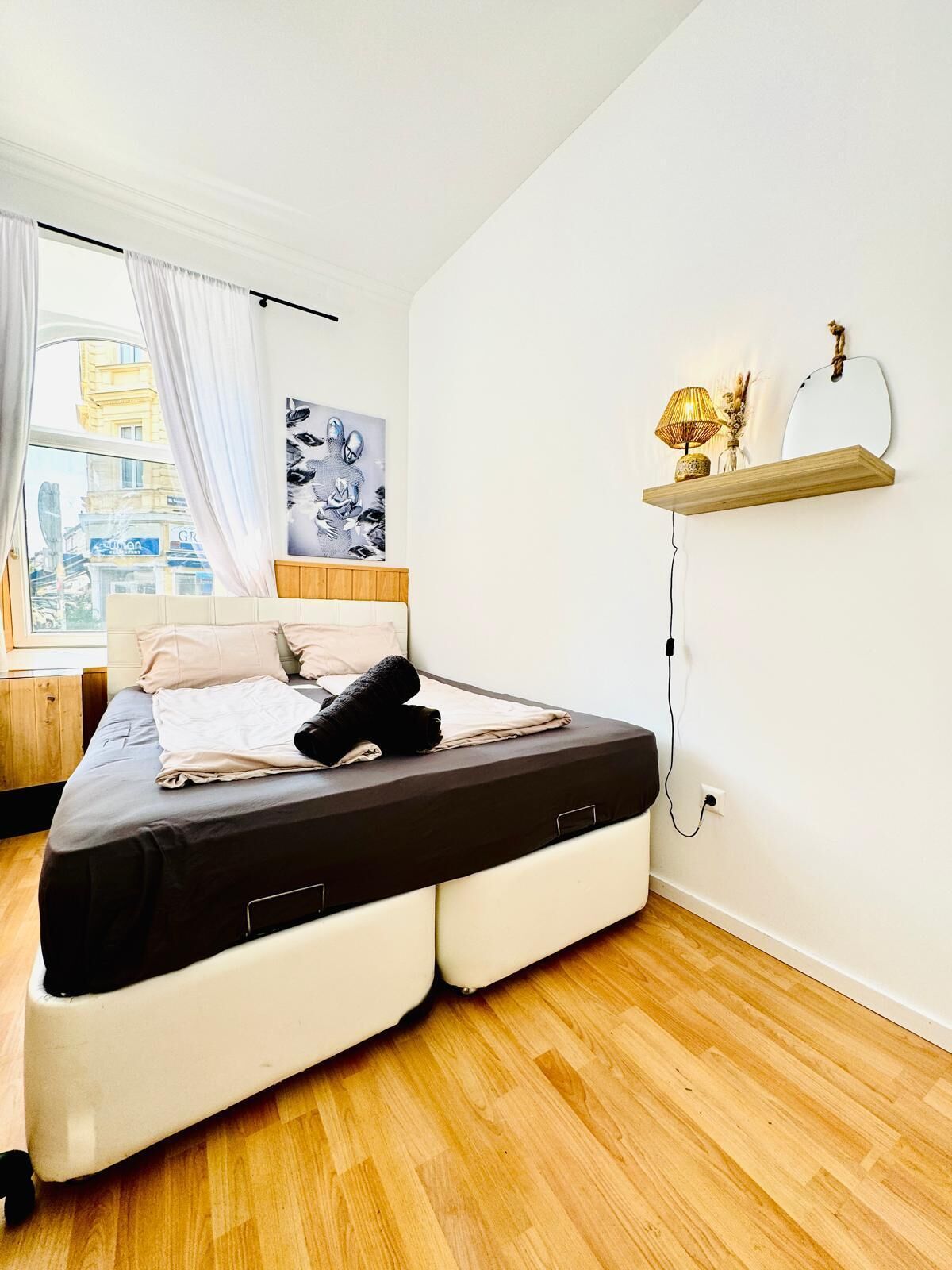 Een moderne slaapkamer met een bed, een raam met gordijnen, een wandplank met een lamp en een schilderij.