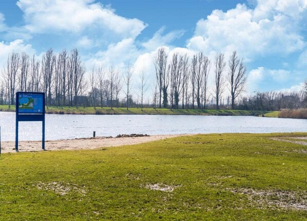 A grassy area with a body of water and leafless trees in the background. - Bungalow in South Holland With Private Jetty (Simonshaven)