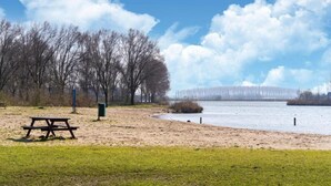 A park with a picnic table and a bench, a sandy area, and a body of water with a bridge in the background. - Bungalow in South Holland With Private Jetty (Simonshaven)