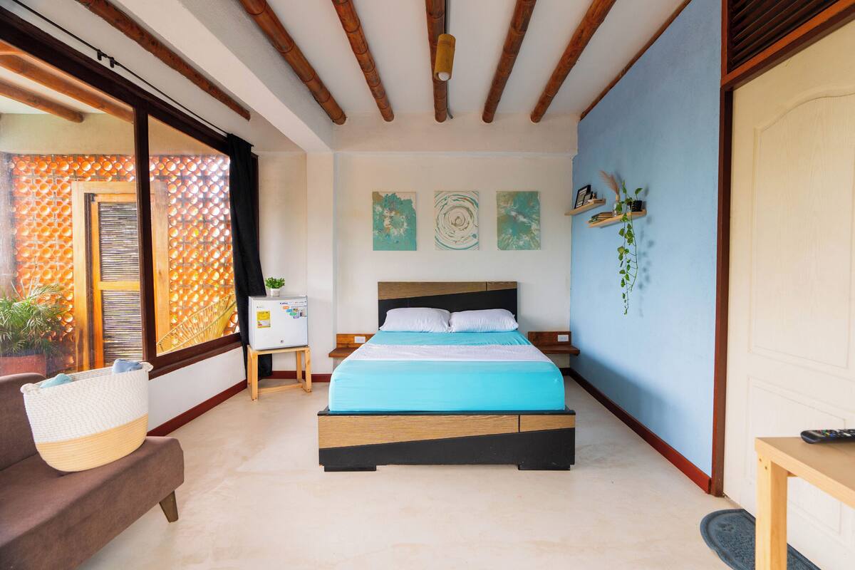 Un dormitorio con cama, cabecera de madera, una mesita de noche, una pequeña mesa y una cesta grande de mimbre.