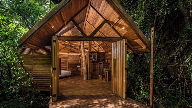 Una cabaña de madera con techo inclinado, una terraza de madera y un dormitorio con cama y escritorio.