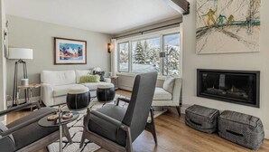 A modern living room with a sofa, armchair, and coffee table, large windows with a view of snow-covered trees, a fireplace, and artwork on the walls.