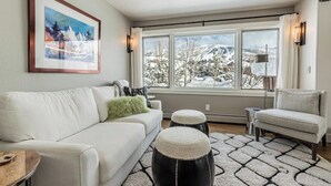 A modern living room with a white sofa, a green throw pillow, a black ottoman, and a view of a snowy landscape through large windows.