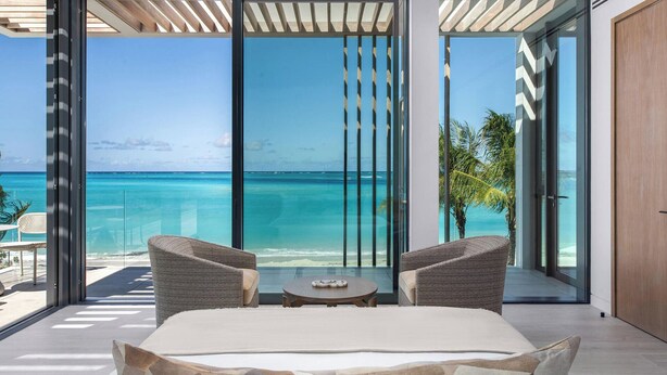 Een slaapkamer met een groot raam met uitzicht op de oceaan. Er staan twee rieten stoelen en een klein rond tafeltje.