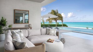 Een moderne woonkamer met een ruime hoekbank, een klein rond voetenbankje en een schaal met lekkernijen. Een groot raam biedt uitzicht op een tropisch strand en de oceaan.