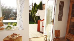 Quarto com piso de madeira, uma pequena mesa de madeira com uma tigela decorativa, janela com cortinas florais e porta que dá para área externa com deck de madeira e vegetação.