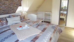 Quarto com teto inclinado, berço, cama com roupa de cama estampada e vista através de uma porta.