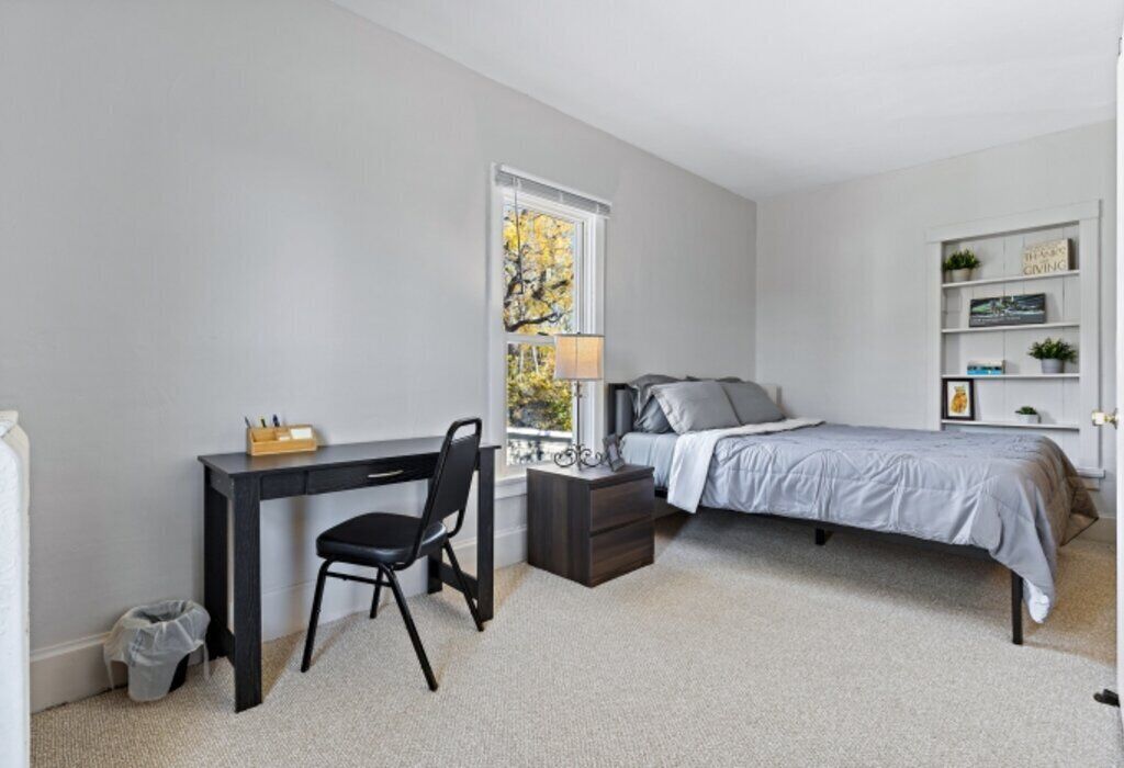 A bedroom with a bed, a desk, a chair, a nightstand, a lamp, and a window.