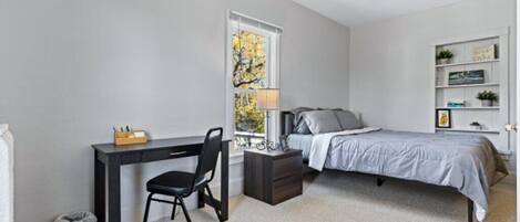 A bedroom with a bed, a desk, a chair, a nightstand, a lamp, and a window.