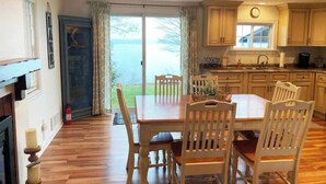 A dining area with a wooden table and chairs, a kitchen with light-colored cabinets, and a view of a body of water through a glass door. - Quiet/charming 2-bedroom Sunset-Lake view cottage lovely Seneca lake, wine trail (Geneva)