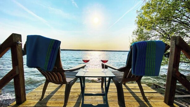 A wooden deck with two chairs and a table, overlooking a body of water. - Quiet/charming 2-bedroom Sunset-Lake view cottage lovely Seneca lake, wine trail (Geneva)