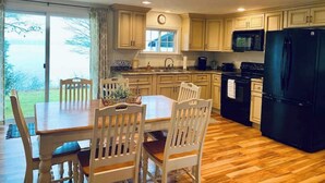 A kitchen with wooden cabinets, a black refrigerator, a dining table with chairs, and a view of the outdoors through a sliding glass door. - Quiet/charming 2-bedroom Sunset-Lake view cottage lovely Seneca lake, wine trail (Geneva)