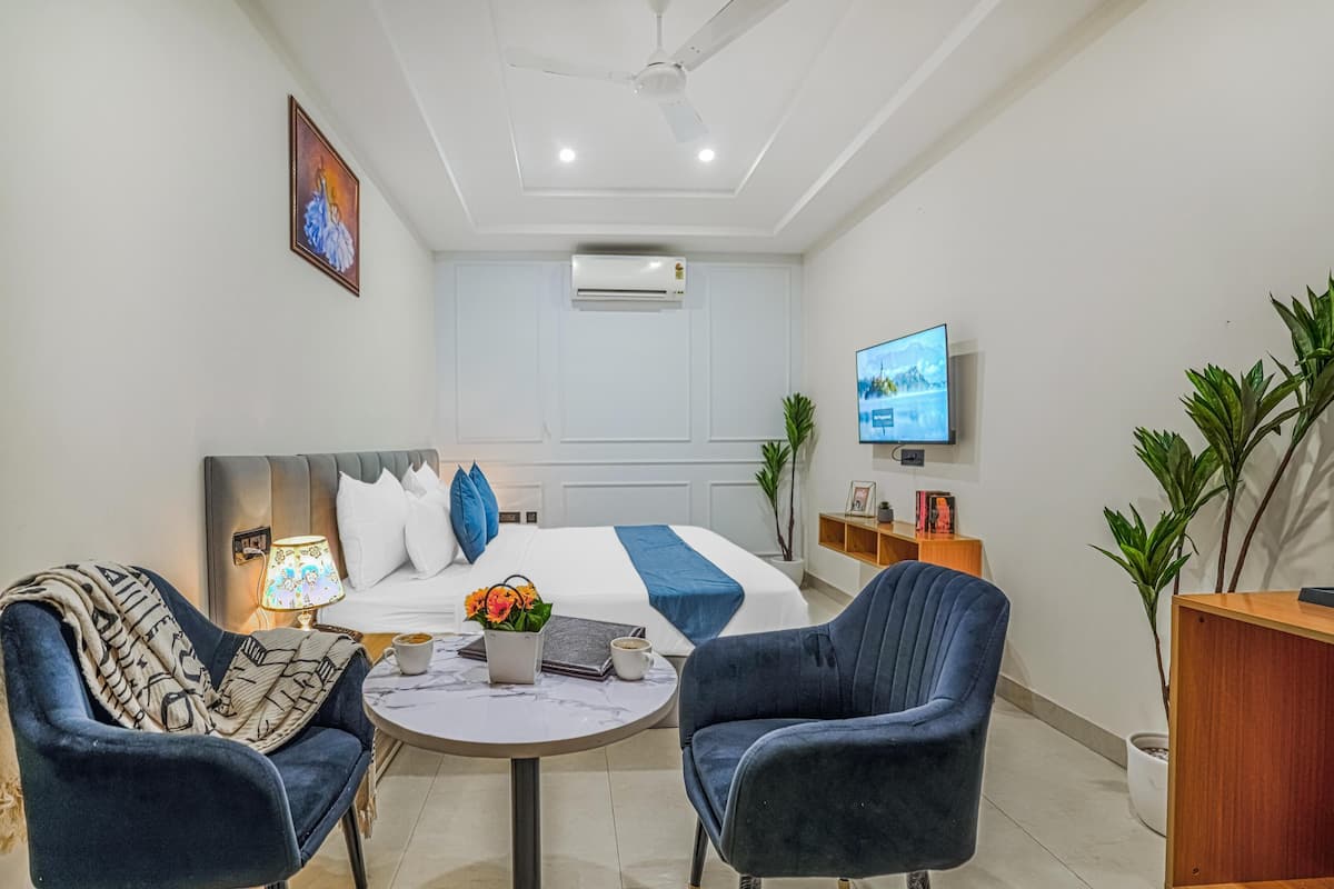 A modern hotel room with a bed, two armchairs, a small table with a lamp, a TV, and a plant.