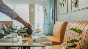 A person pouring coffee into a cup at a table with a plant and a sofa in the background. - Hotel Sky View - Zirakpur, Chandigarh (Dera Bassi)