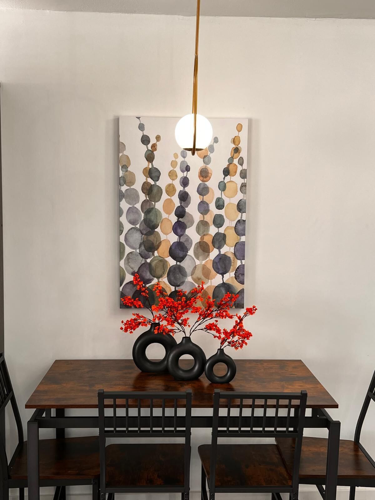 A dining area with a wooden table and chairs, a hanging light fixture, and a wall-mounted artwork with abstract circles and red flowers.