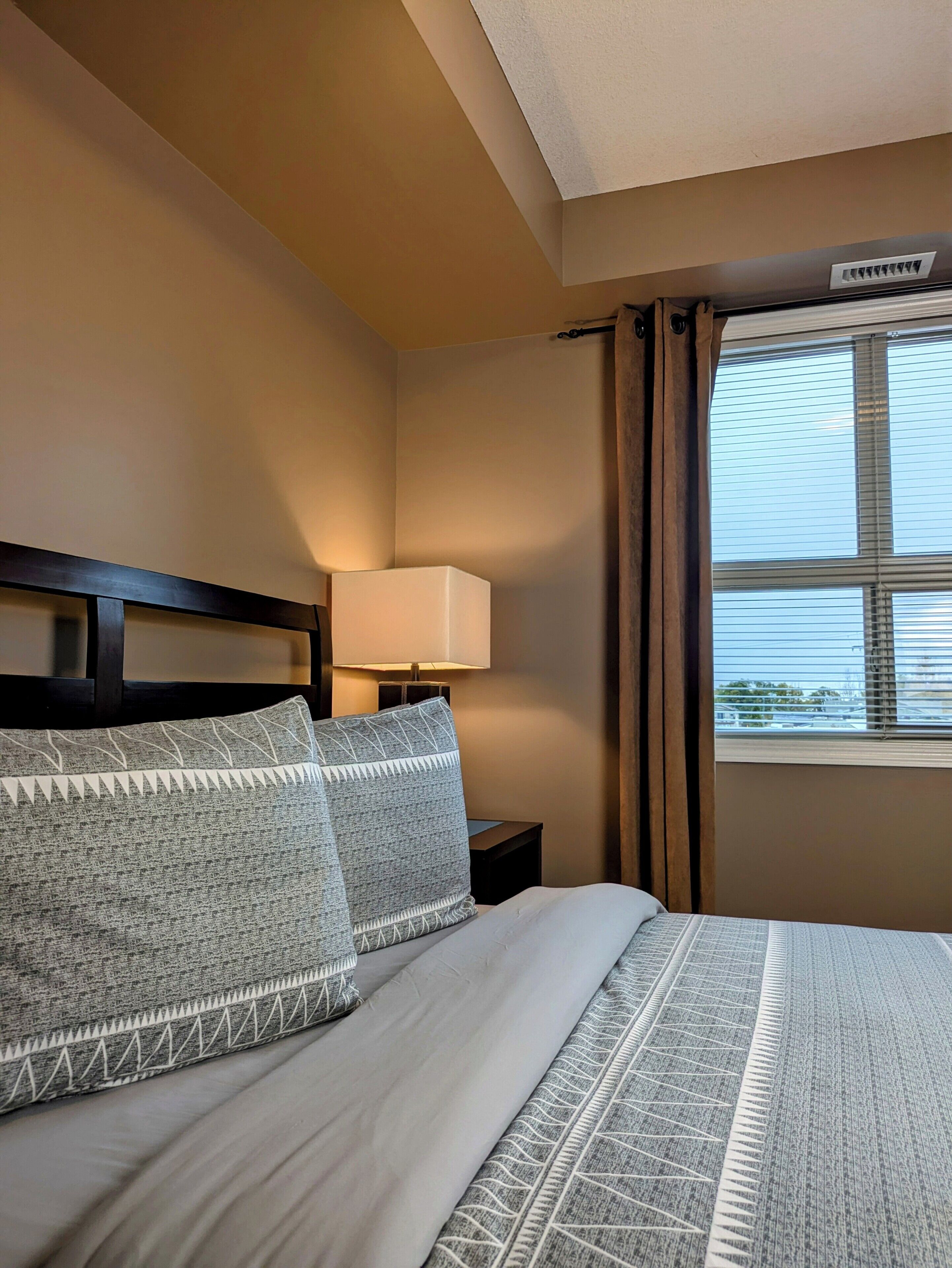 A hotel room with a bed, a nightstand with a lamp, a window with curtains, and a ceiling vent.