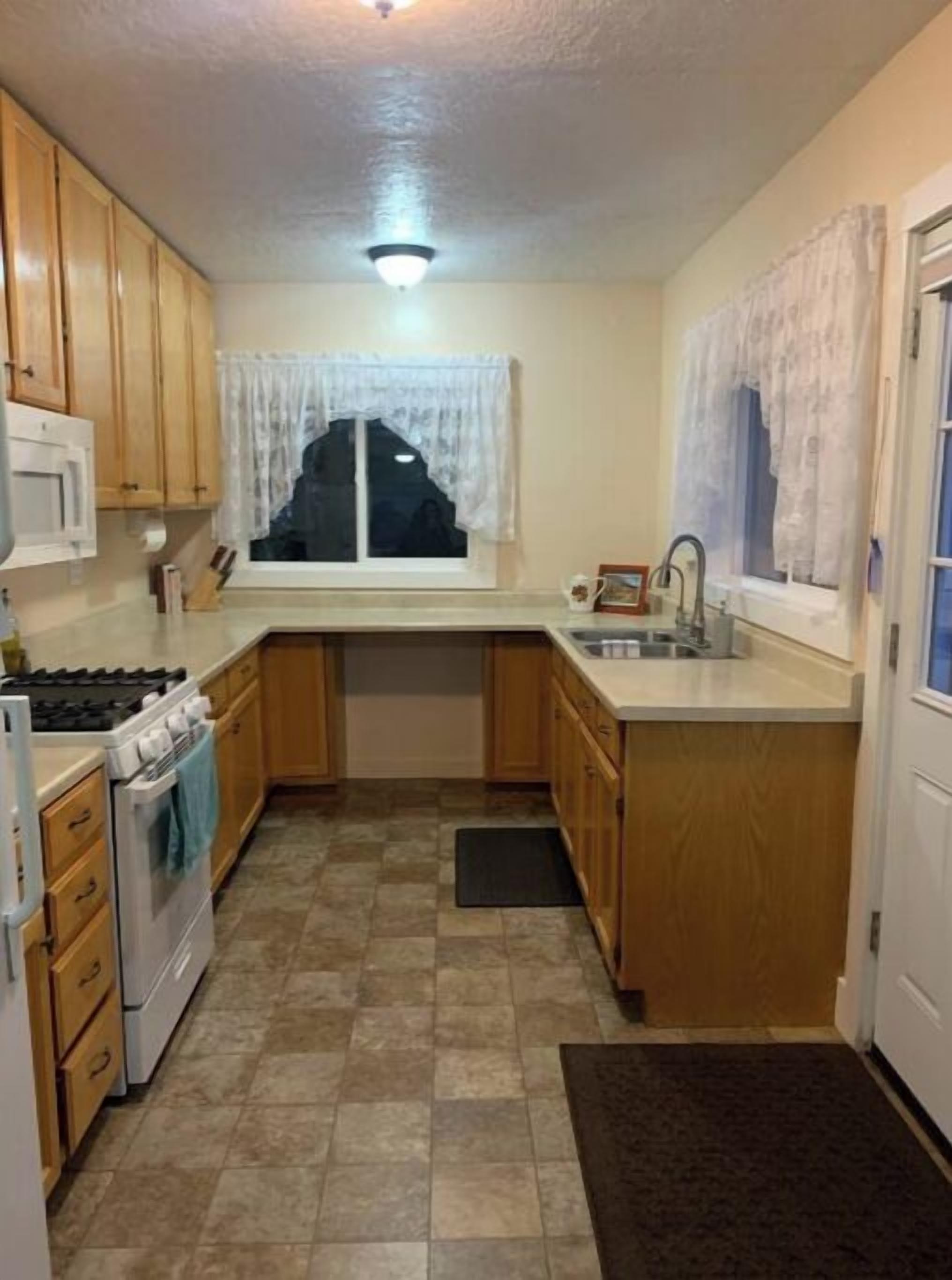 A compact kitchen with wooden cabinets, a white stove, a sink, and a window with white curtains.