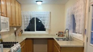 A compact kitchen with wooden cabinets, a white stove, a sink, and a window with white curtains. - The Guesthouse (Great Falls)