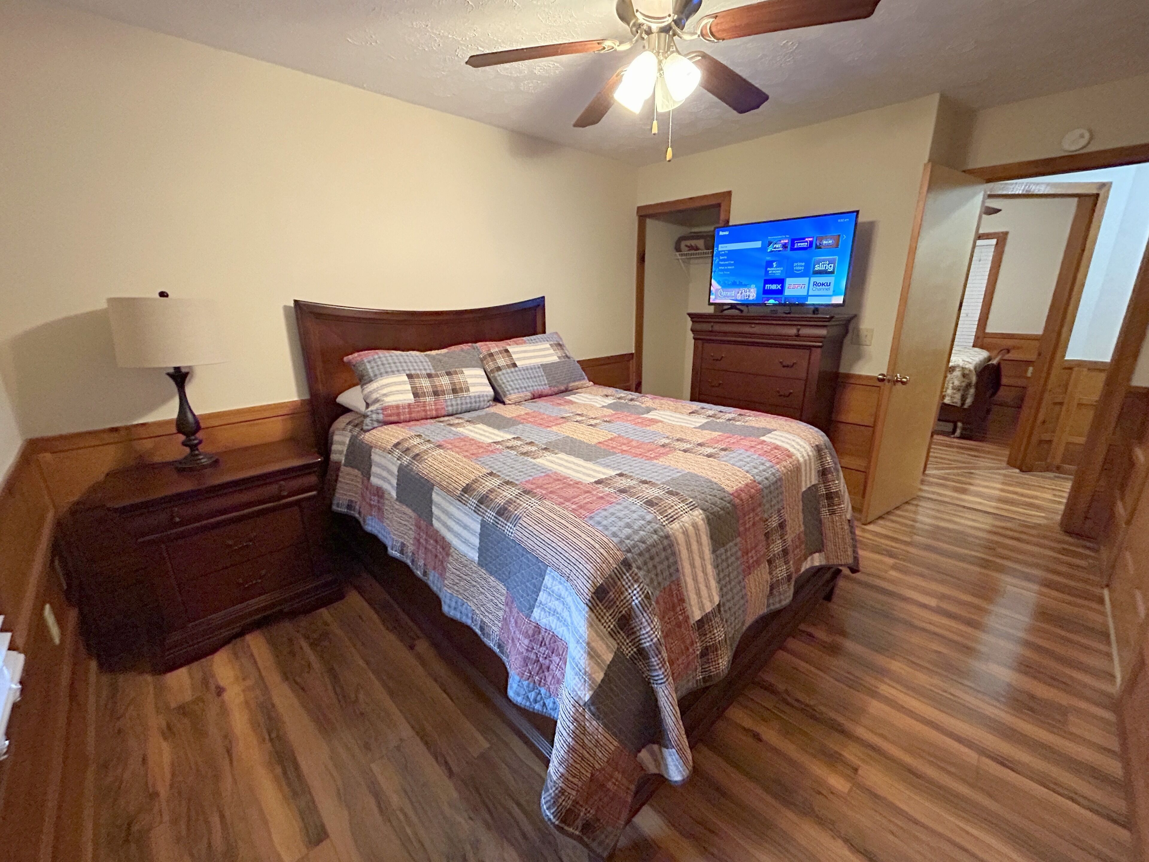 A bedroom with a wooden floor, a bed with a quilt, a nightstand, a lamp, a dresser, a television, and a ceiling fan.