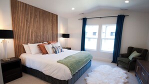 Een moderne slaapkamer met een groot bed, een houten accentmuur, een nachtkastje met lamp, een stoel, een dressoir en een groot raam met gordijnen.
