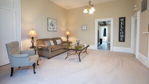 Un salon avec un tapis beige, un fauteuil bleu clair, un canapé marron, deux lampes, une table basse et un panneau « BIENVENUE » accroché au mur.