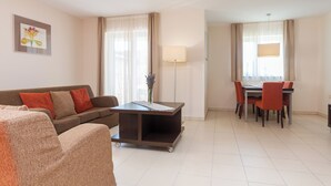 A hotel room with a sofa, a small table with a lamp, a dining table with chairs, and a large window with curtains.
