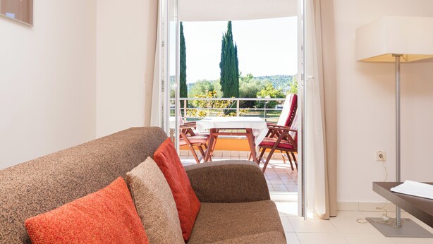 A room with a sofa, a table, and chairs on a balcony with a view of trees and a distant hill.