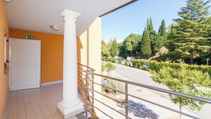 A balcony with a white door, a metal railing, and a view of a road with trees and parked cars.