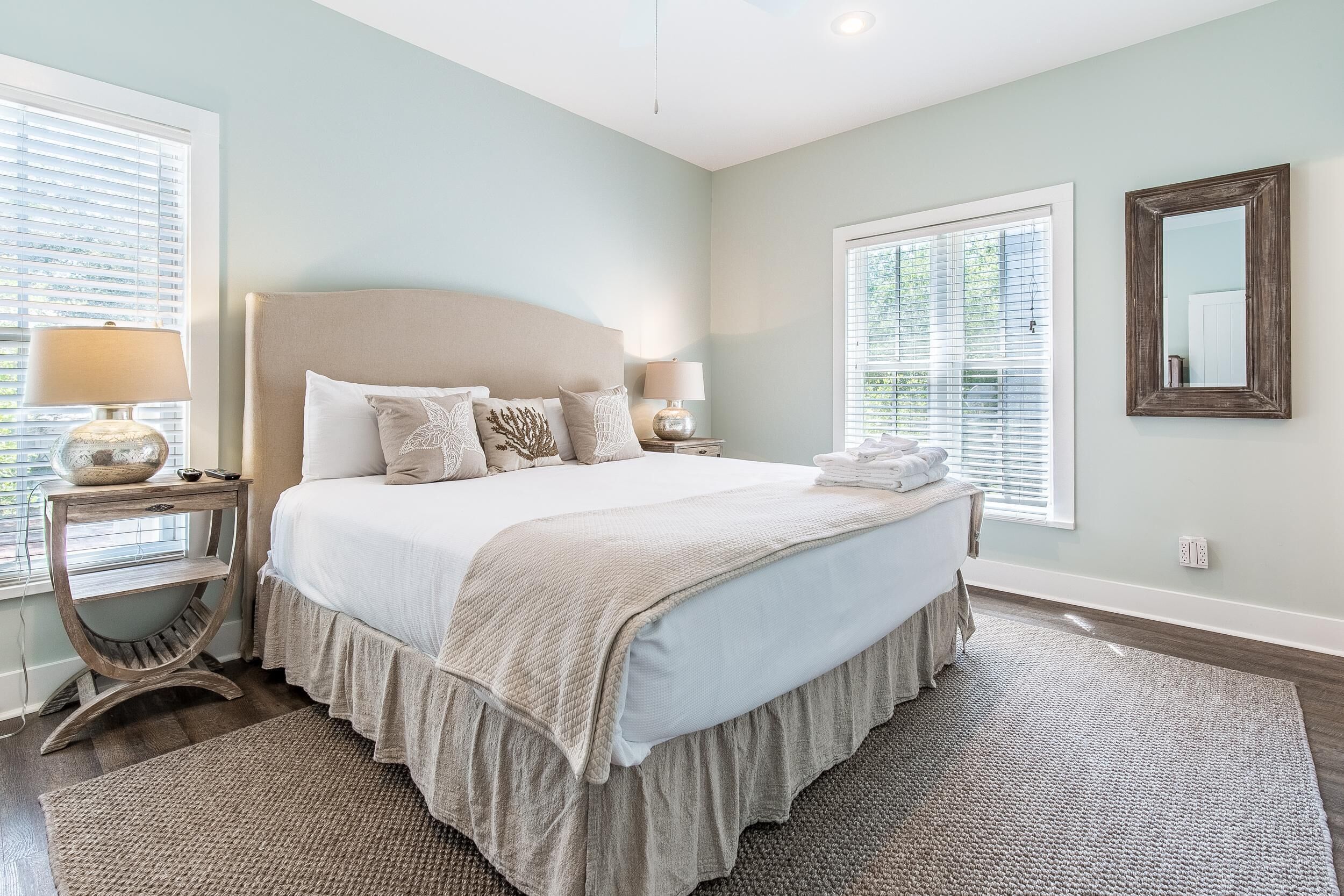 Une chambre avec un grand lit, deux tables de chevet, une fenêtre avec des stores, un miroir et un tapis.