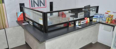 Un mostrador de recepción de hotel con un separador de vidrio, un letrero que dice 'KT INN' y varios objetos sobre el mostrador, incluyendo un dispensador de agua y aperitivos.