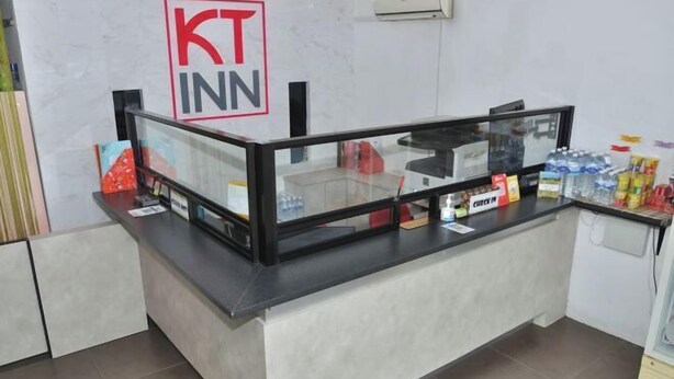 A check-in counter with a glass partition, a sign reading "KT INN," and various items like water bottles and snacks on the counter. - Kuching Transit Inn (Kuching)