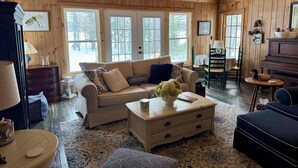 A cozy cabin interior with a wooden ceiling, large windows, a sofa, a coffee table, and a piano.
