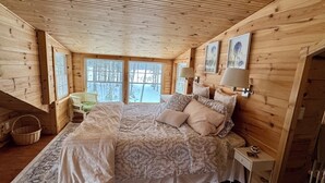 A cozy wooden cabin bedroom with a large bed, a nightstand, a lamp, and a view of the snowy landscape through the windows.