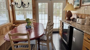 A cozy cabin interior with a wooden dining table, chairs, a chandelier, a kitchen with a refrigerator, and a large window with a view of a snowy landscape.