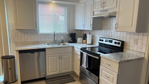 A modern kitchen with white cabinetry, a stainless steel dishwasher, a black stove, and a window with blinds.