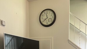 A living room with a flat-screen TV on a white entertainment unit, a clock on the wall, and a staircase to the right.