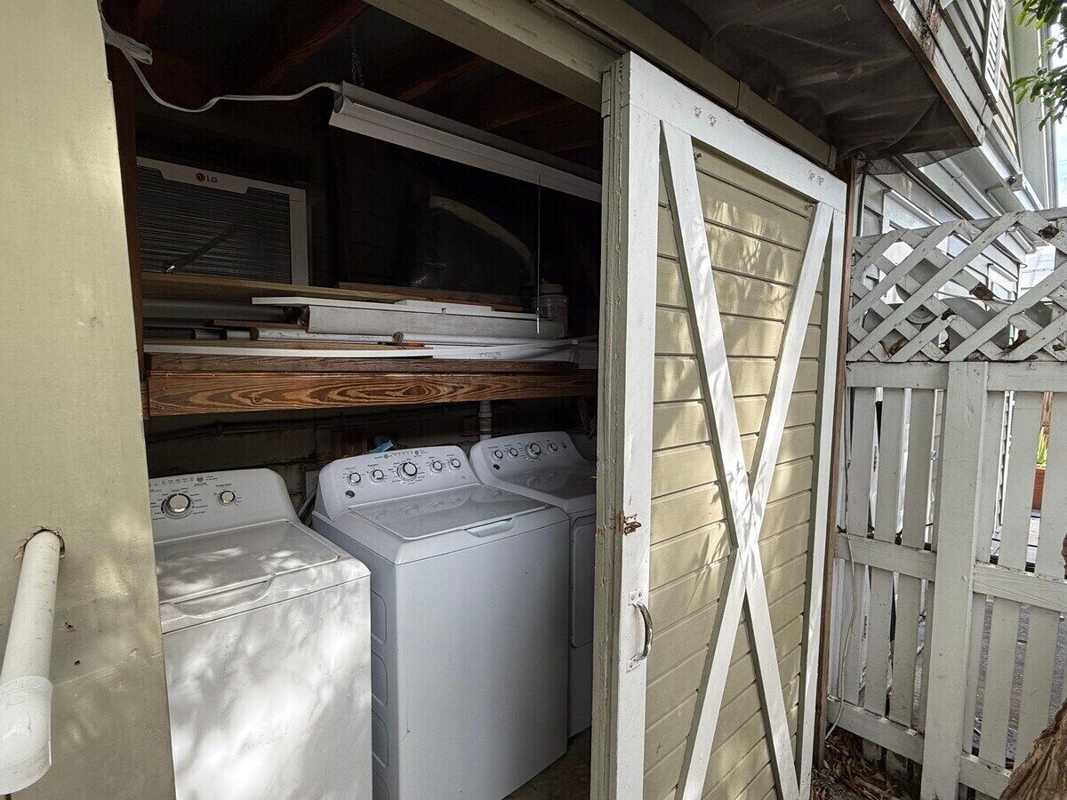 Un área de lavandería con dos lavadoras y una secadora, un estante de madera encima de las máquinas, y una cerca blanca en el exterior.