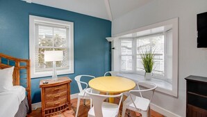 A room with a bed, a round yellow table with white chairs, a wicker side table with a lamp, and a window with blinds. - Ibis Suite | Bright Hideaway, Hot Tubs, Steps from Duval! (Key West)