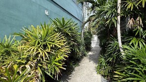 A narrow concrete pathway flanked by lush green plants and a blue wall. - Ibis Suite | Bright Hideaway, Hot Tubs, Steps from Duval! (Key West)