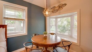 A dining area with a round wooden table, four chairs, a hanging light fixture, and two windows with blinds. - Pelican Suite | Cozy Cottage, Hot Tubs, Close to D (Key West)