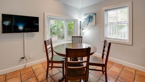 Un coin repas avec une table ronde, quatre chaises en bois, un téléviseur à écran plat fixé au mur et deux fenêtres équipées de stores.