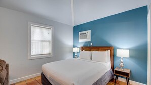 Une chambre à coucher avec un lit, une table de chevet avec une lampe, une fenêtre avec des stores et un climatiseur fixé au mur.
