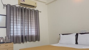 Ett hotellrum med en säng, ett litet bord vid sängen med en vattenkokare, ett litet skåp och ett fönster med gardiner.