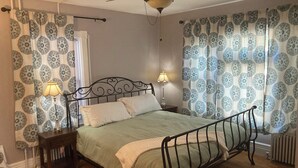 Un dormitorio con estructura de cama metálica, dos lámparas de noche, cortinas estampadas y ventilador de techo.