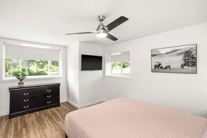 Une chambre à coucher comprenant un lit, une commode, un ventilateur de plafond, un téléviseur et une fenêtre donnant sur des arbres.