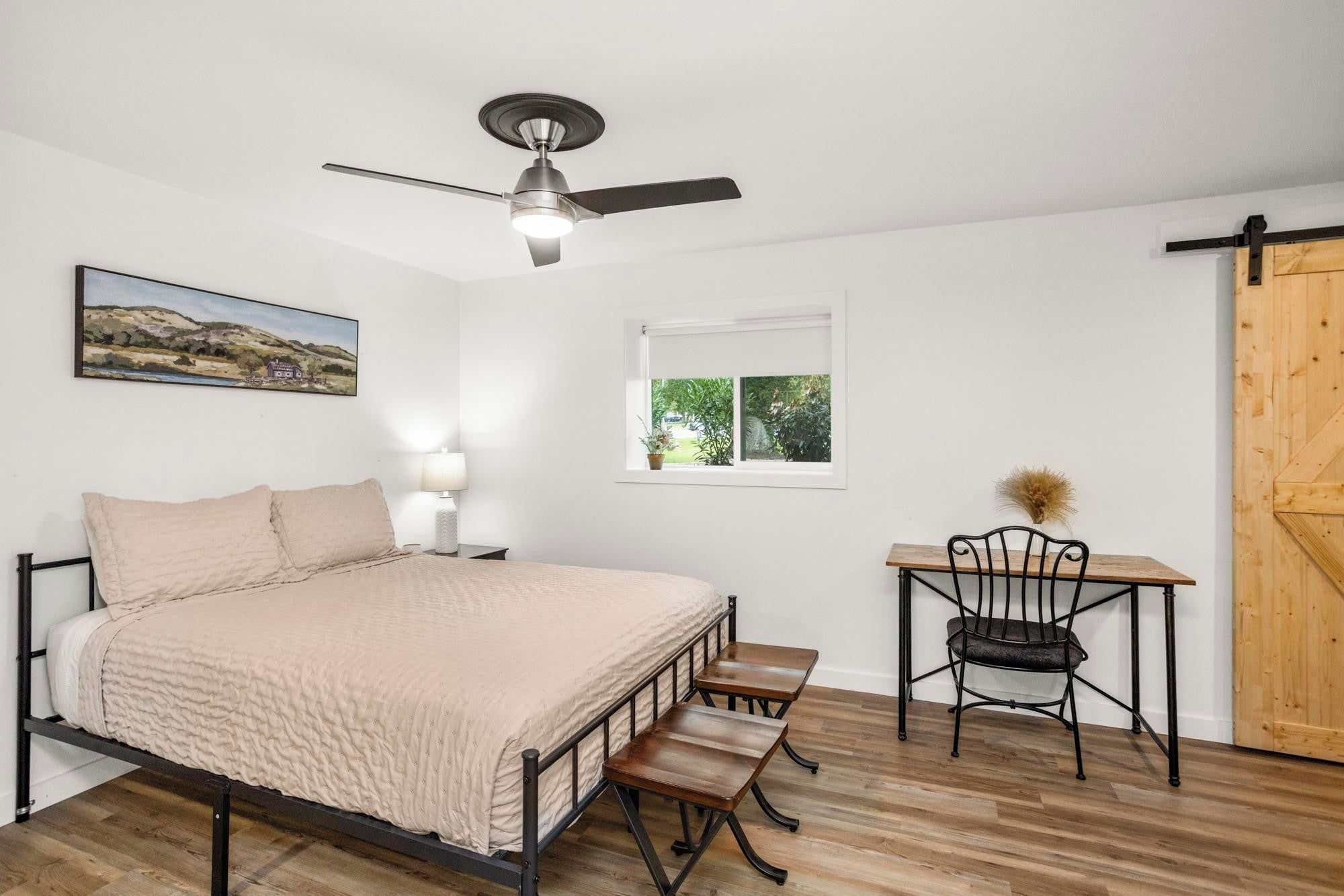 A bedroom with a bed, a desk, a chair, a ceiling fan, a window, and a door.