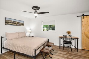 A bedroom with a bed, a desk, a chair, a ceiling fan, a window, and a door.
